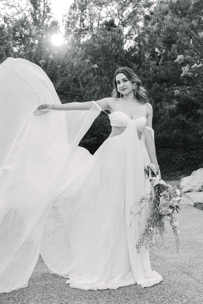 A woman poses gracefully outdoors in a flowing, sleeveless white dress, holding a bouquet of flowers. The sun peeks through the trees behind her, casting a gentle light on the scene. The image has a serene, natural ambiance.
