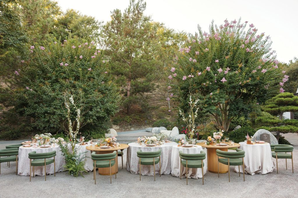 An elegant outdoor dining setup features two round tables with white tablecloths and green chairs. The tables are adorned with floral arrangements and serveware. Tall trees in the background add a natural ambiance to the scene.