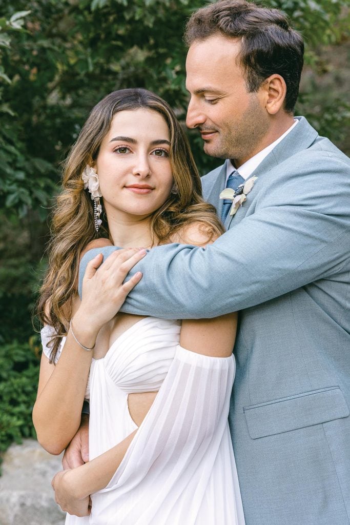A couple embraces outdoors. The woman, in a flowing white dress, stands with a gentle smile. The man, in a light blue suit, lovingly wraps his arm around her shoulder. Green foliage provides a natural background.