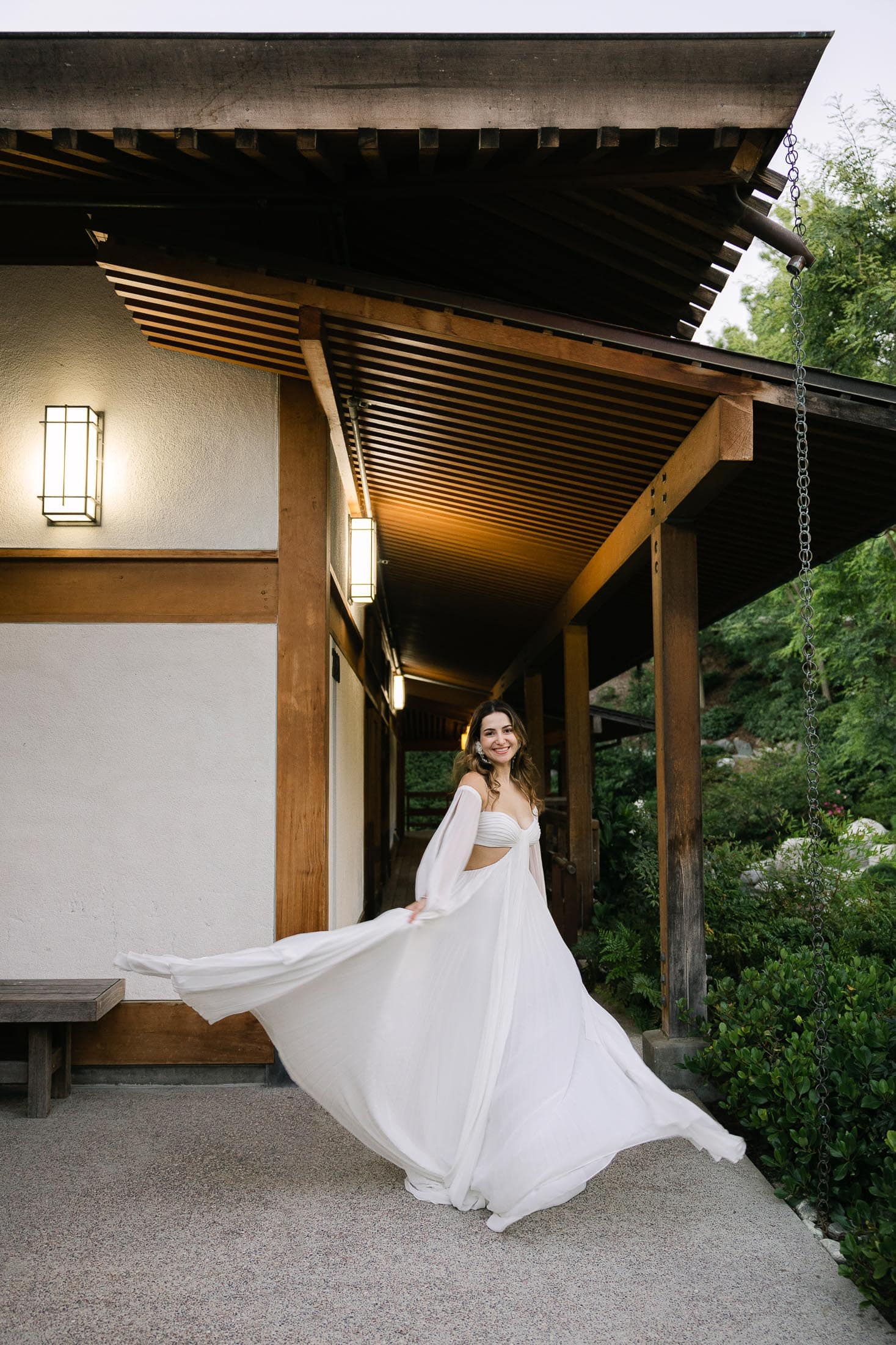 A person in a flowing white dress twirls joyfully in front of a building with wooden beams and a slanted roof at the Japanese Friendship Garden. The dress billows gracefully as they move under the warm light of lanterns.