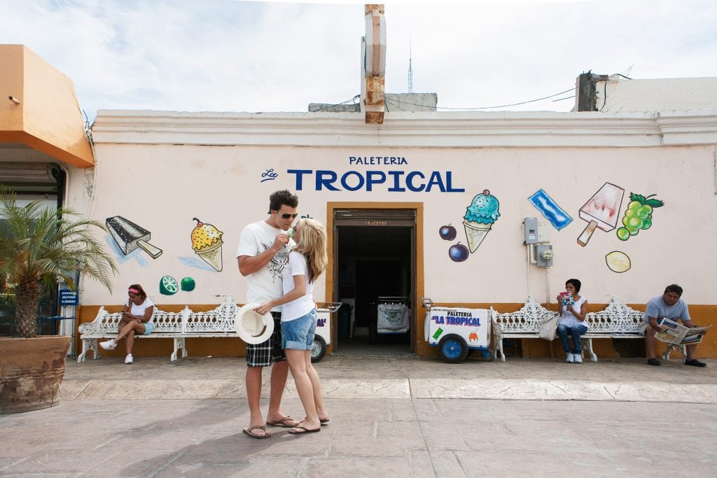 A couple stands in front of a colorful ice cream shop named "La Tropical" in San Diego, sharing a moment perfect for engagement photos. The shop's walls boast vibrant ice cream illustrations. Nearby, several people enjoy the sunny day from benches.