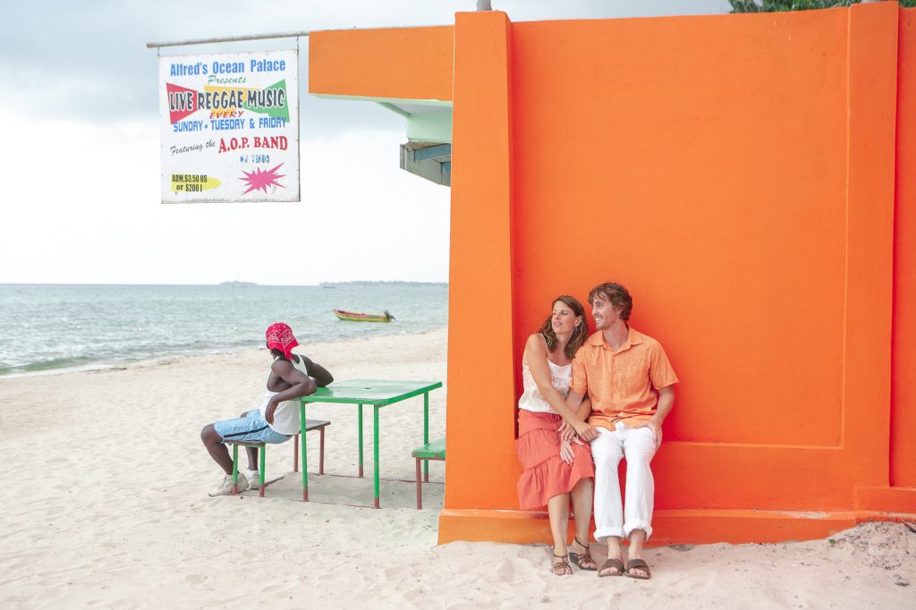 A couple sits on the steps of a bright orange building at a beach, smiling and talking. Nearby, a person in a red hat sits at a green table as live reggae music is advertised. The ocean and a boat are visible in the background, perfectly capturing San Diego engagement photos' charm.