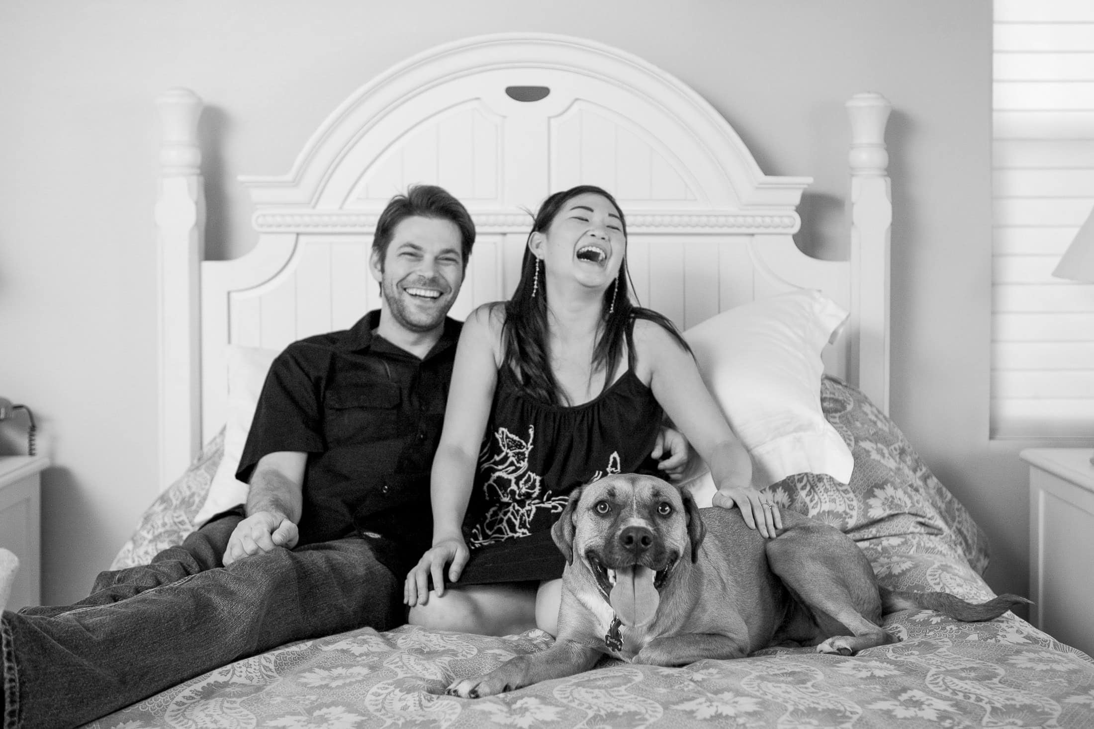 A black and white image captures the essence of San Diego engagement photos— a couple sitting on a bed with their large dog, laughing and relaxed. The dog sits contently between them in the airy room, complete with a white headboard, embodying joy and tranquility.
