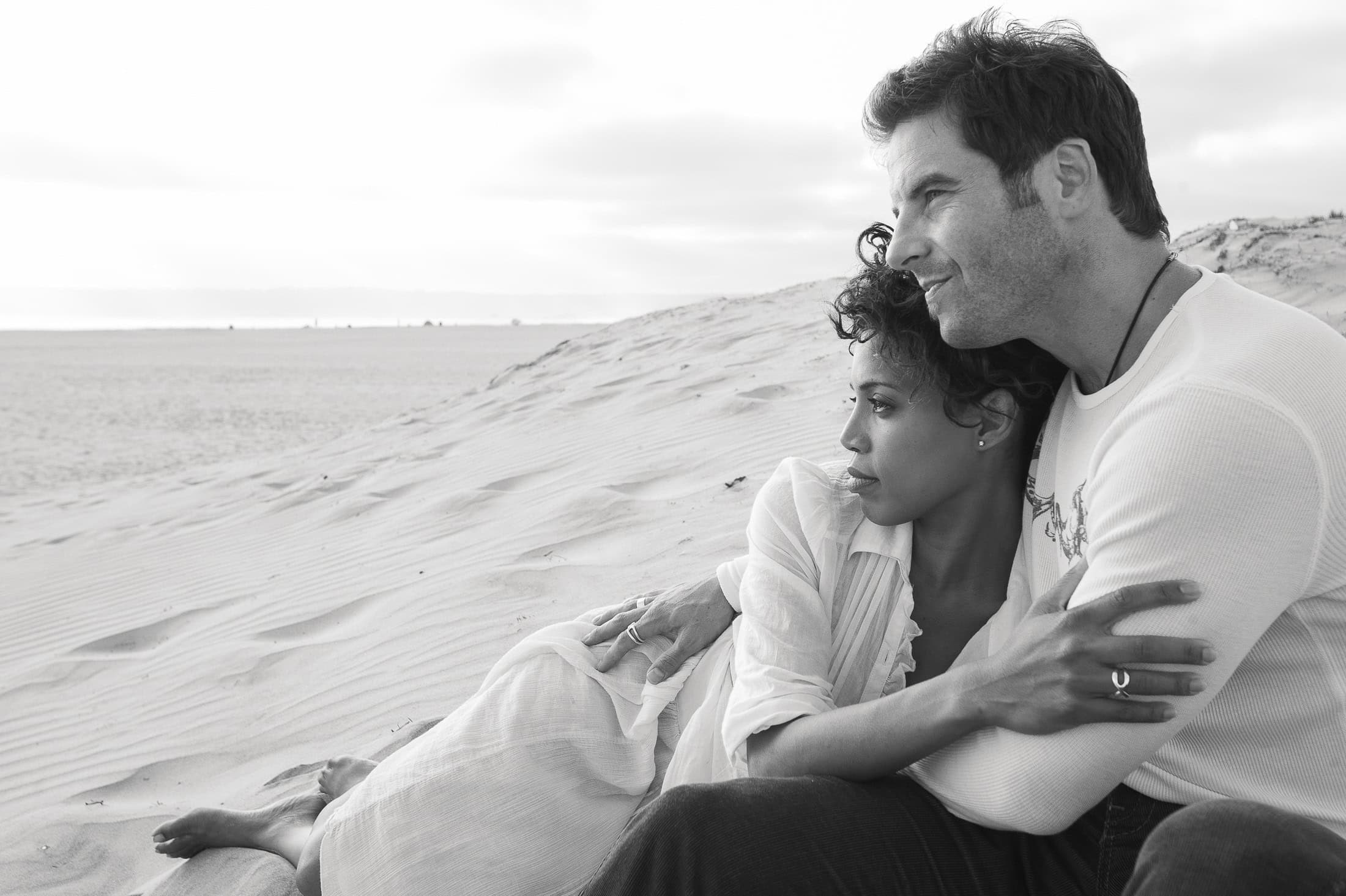 A couple sits together on a sandy beach, embracing while gazing at the horizon. The woman leans on the man's chest, their San Diego engagement photos capturing this timeless, serene scene in black and white.