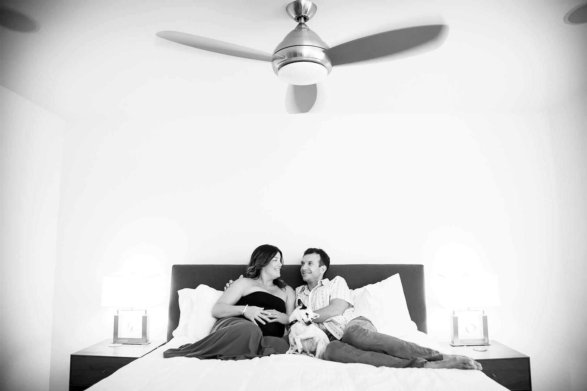 A couple relaxes on a bed, smiling at each other, with a small dog resting between them. The room has a modern ceiling fan above, two lit lamps on nightstands, and white bedding. The scene is in black and white.
