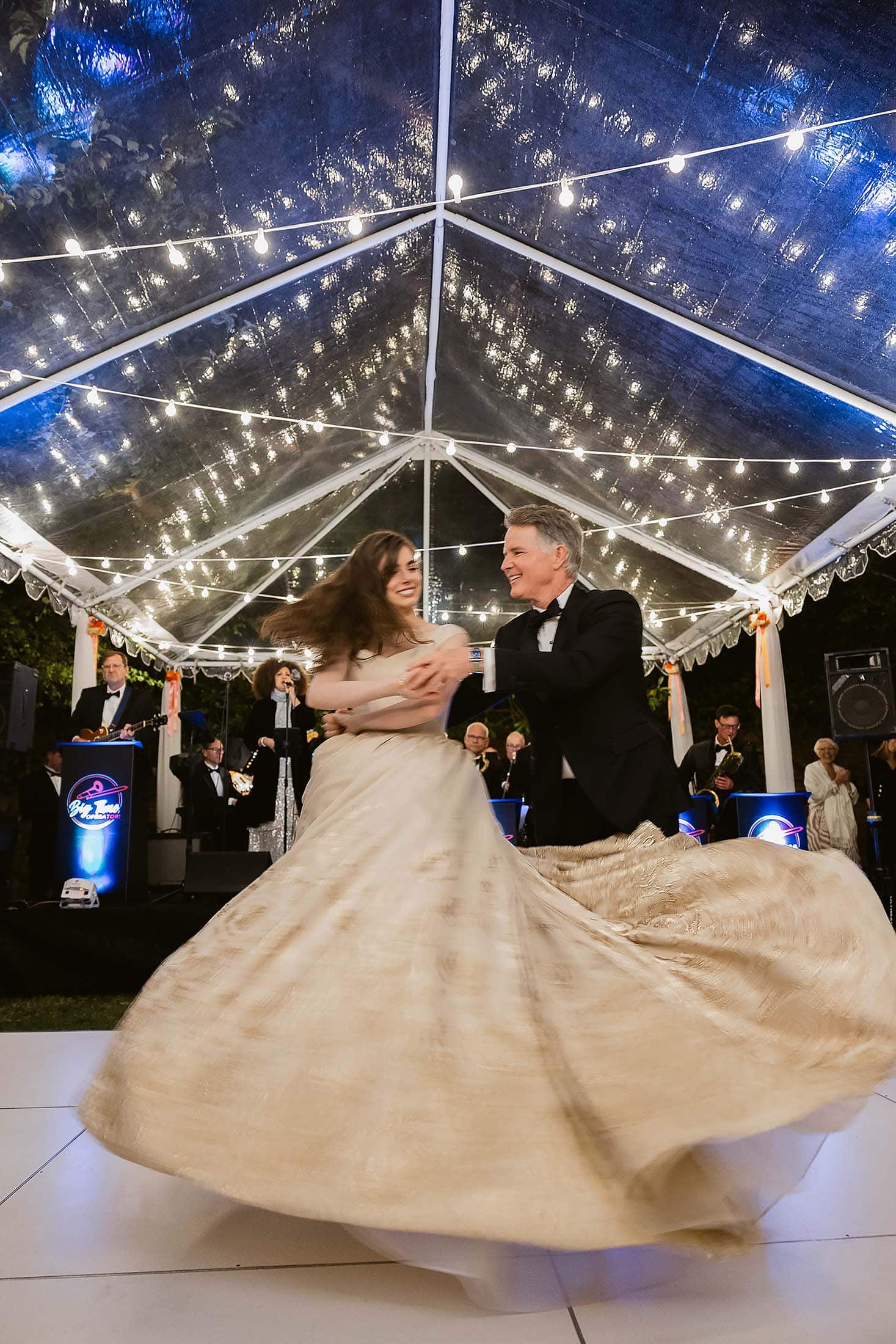Father Daughter in formal attire dances joyfully under a clear tent with string lights, surrounded by guests and a live band. The woman's beige gown swirls dramatically as they spin together—it's all about celebration on the glowing dance floor.