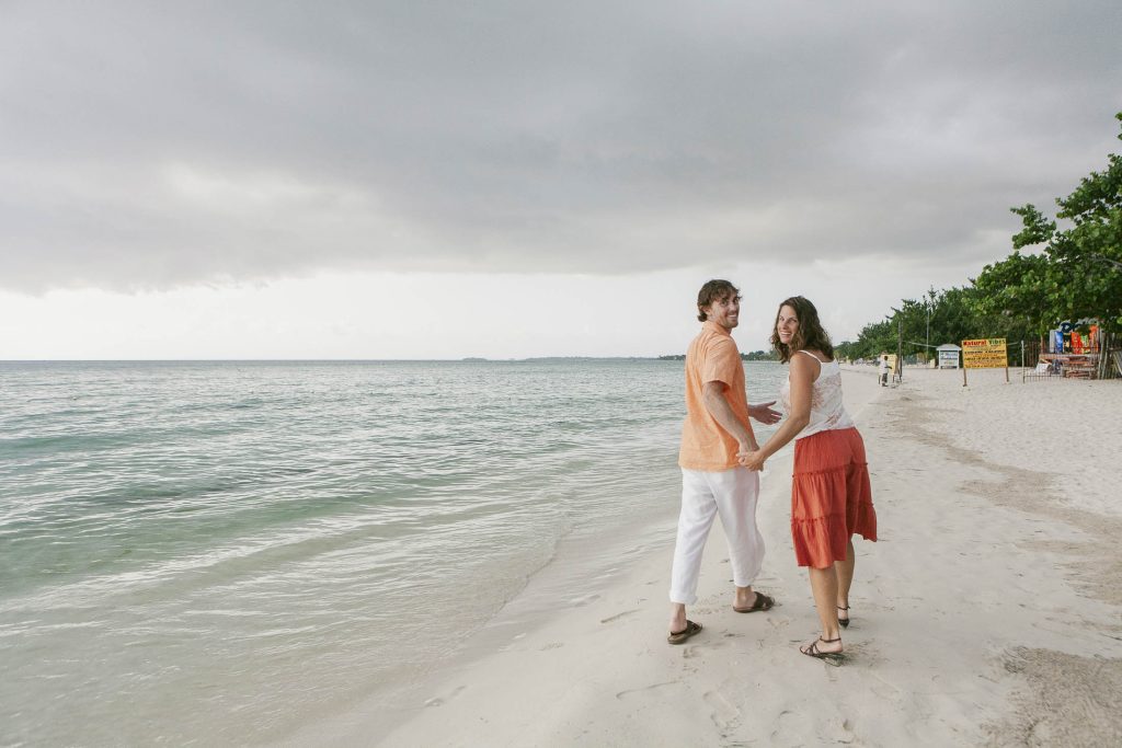 A smiling couple walks hand-in-hand along a sandy beach, looking back toward the camera. The ocean is calm and the sky is cloudy, with trees and signs visible in the background.