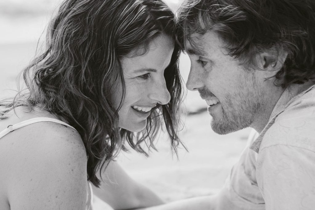 A black and white photo of a smiling couple facing each other closely, touching foreheads and looking into each other's eyes, sharing an intimate moment.