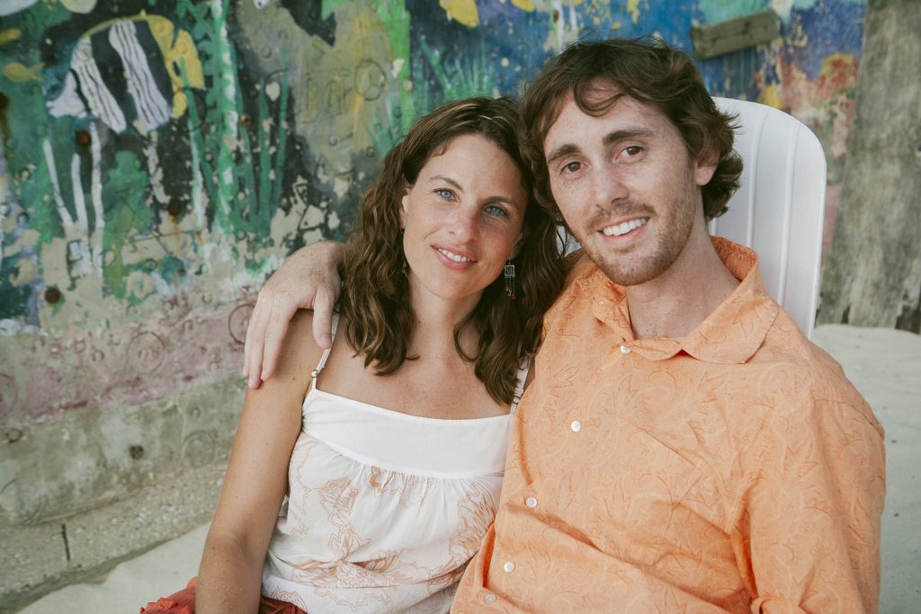 A smiling couple sits close together outdoors, with the man’s arm around the woman. They are seated in front of a colorful, abstract mural. Both look relaxed and happy.