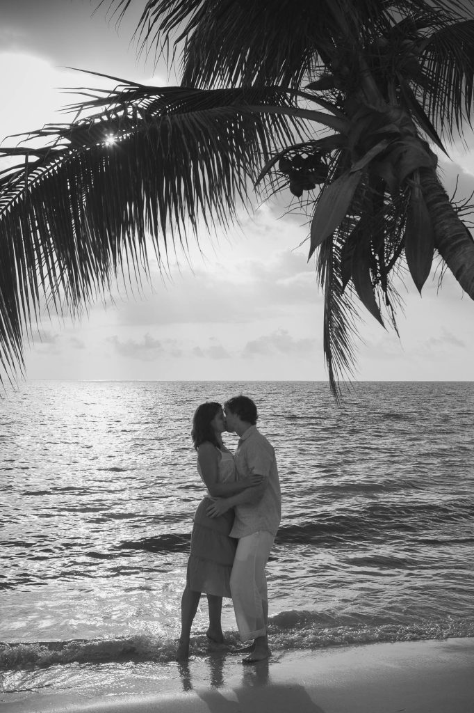 Negril, Jamaica: A couple stands on the beach at sunset, embracing and sharing a kiss beneath a palm tree, with the ocean and a bright sky in the background. The image is in black and white.