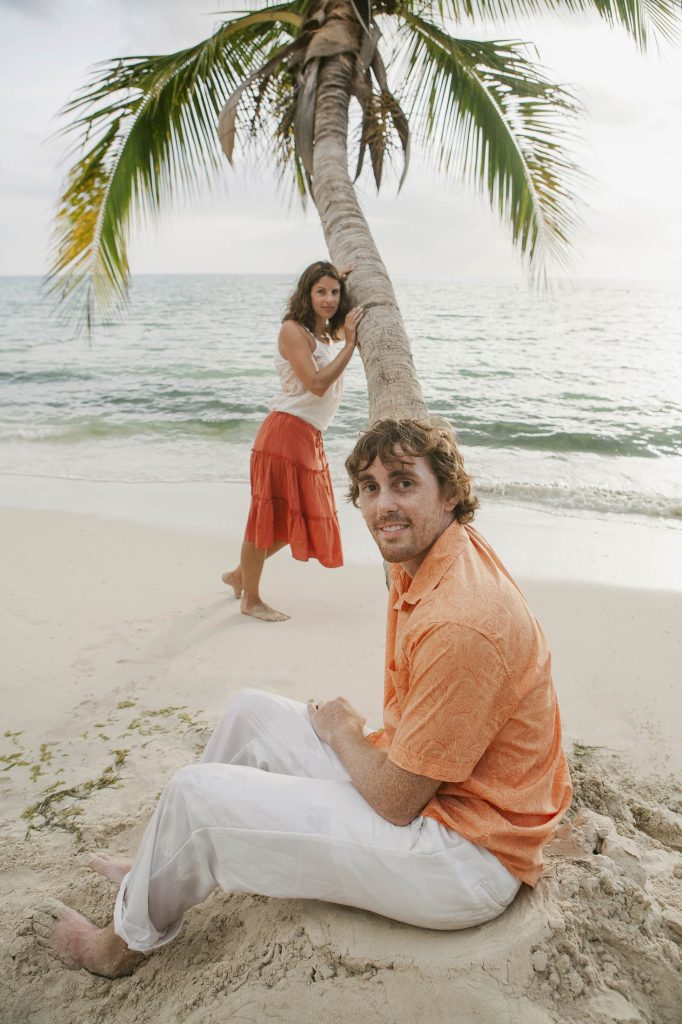 Negril, Jamaica: A man in an orange shirt sits on sandy beach, smiling at the camera, while a woman in a red skirt and white top stands behind him, leaning against a palm tree by the ocean.