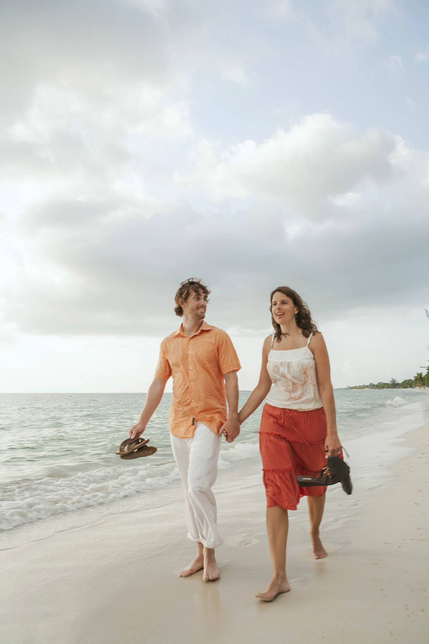 Negril, Jamaica: A couple walks barefoot on a sandy beach, holding hands and their shoes, with the ocean and cloudy sky in the background. They are smiling and enjoying the peaceful seaside.