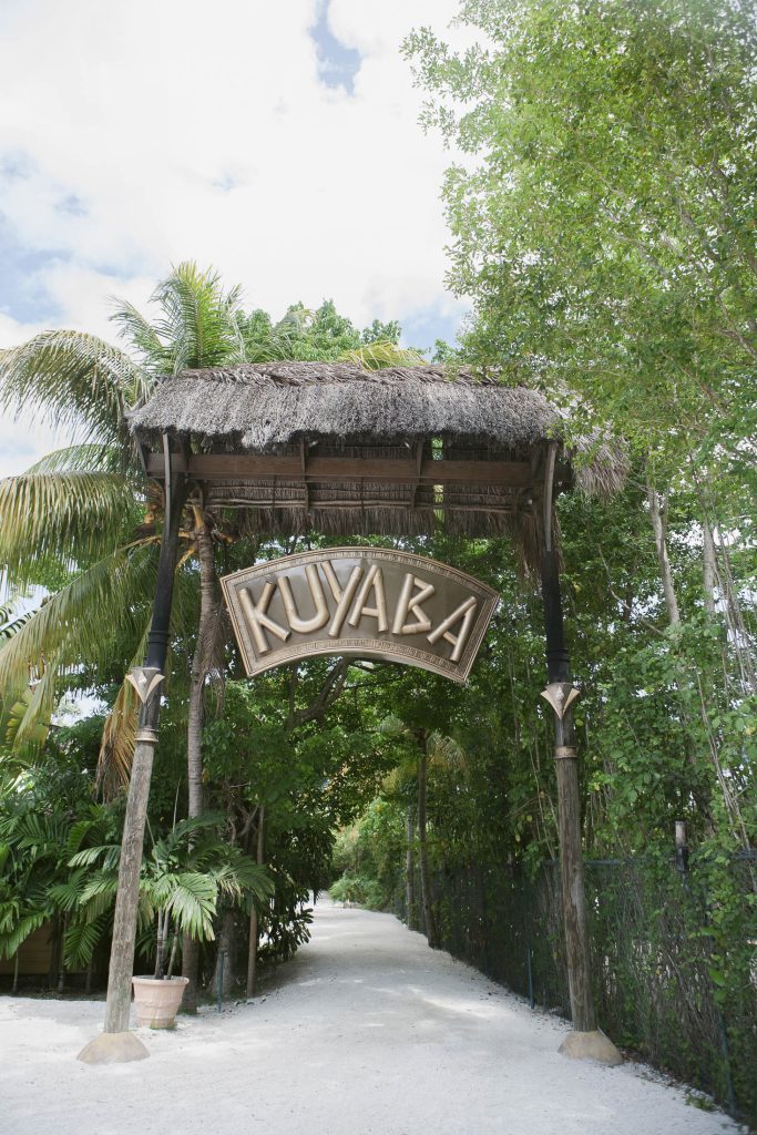 Negril, Jamaica: A wooden archway with a thatched roof and a sign reading "KUYABA" stands over a sandy path surrounded by lush green trees and tropical plants.