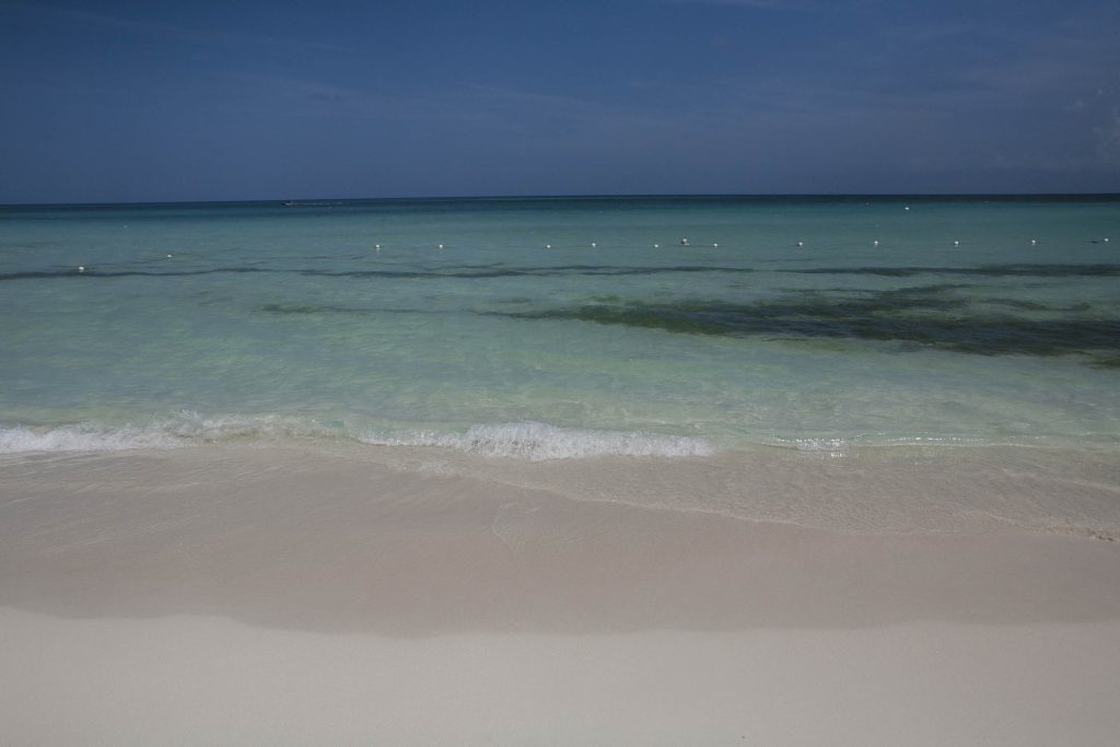 Negril, Jamaica: A tranquil beach scene with gentle waves lapping onto soft, white sand. The clear turquoise water gradually deepens to a darker blue toward the horizon under a bright, clear sky.