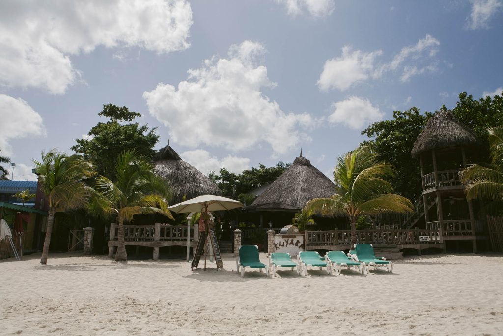 Negril, Jamaica: Four turquoise lounge chairs and a large umbrella sit on a sandy beach, with palm trees and several thatched-roof huts in the background under a partly cloudy sky.
